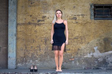 Çekici, orta yaşlı, kısa, siyah, seksi bir elbise giyen, ayakkabıları yanında düz duran, kahverengi bir duvarın önünde nötr bir bakışla duran sarışın kadın.