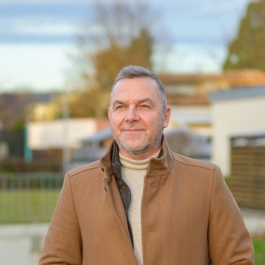 Close up waist up portrait of a middle aged man in a brown coat looking to the side with a gentle smile against an urban background