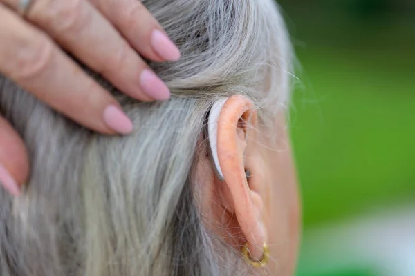 Elderly gray haired hard of hearing lady in her 80\'s brushes her hair to the side to show her hearing aid in her ear