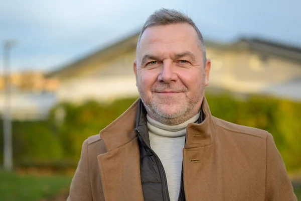 Portrait of a middle aged man in a brown coat smiling softly at the camera against an urban background
