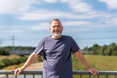 Smiling middle aged man is standing on a bridge with his hands on the railing and looking to the camera