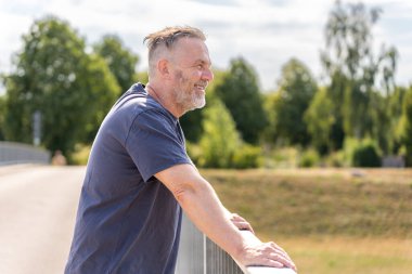 Elderly friendly man is standing on a bridge with his hands on the railing and blue sky looking away