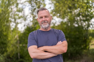 Confident middle aged man standing in front of green trees with crossed arms