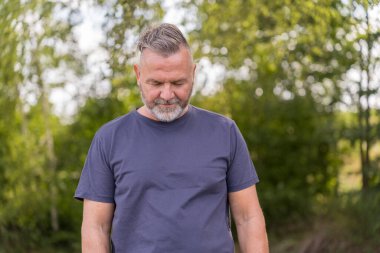 Pensive man in casual attire in front green trees looking down to the ground