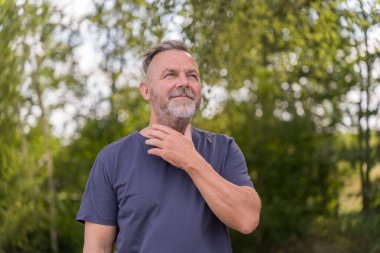 Pensive middle aged man standing in front of green trees with his hand to his neck or throat