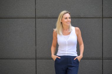 Attractive trendy blond middle aged woman in summer fashion posing against a grey wall with copyspace