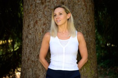 Cute thoughtful trendy blond middle aged woman posing against a tree looking aside with a smile