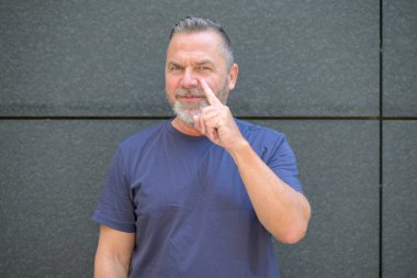 Orta yaşlı sakallı bir adam koyu gri bir duvara karşı poz verirken sol gözünü işaret ediyor.