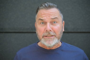 Yaşlı adam panikle kameraya bakıyor. Gri bir duvara karşı geniş gözlü bir ifadeyle.