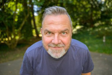 Gülümseyen orta yaşlı bir adam açık havada kesilmiş bir yüz portresiyle kameraya doğru eğiliyor.