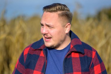 Arka plan olarak bir sonbahar mısır tarlasının önünde kırmızı ve mavi oduncu ceketli ve mavi tişörtlü genç bir adam.