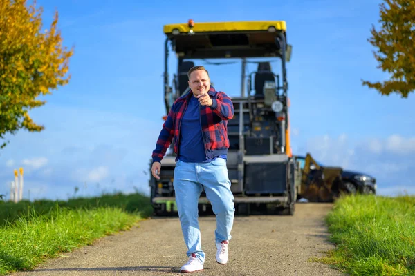 身穿红蓝相间的伐木工夹克和蓝色T恤 面带微笑的年轻人指着摄像机 背对着镜头 后面是农业机械 — 图库照片