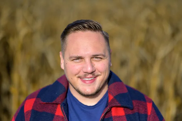 Frontal portrait of a young man with a friendly smile wearing a red and blue lumberjack jacket and blue t-shirt against an autumnal cornfield as a background