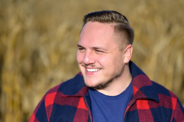 Side portrait of a friendly smiling young man wearing a red and blue lumberjack jacket and blue t-shirt in front of an autumnal corn field as a background
