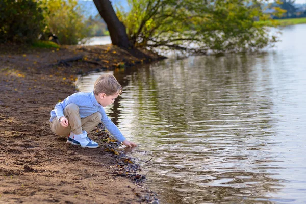 stock image Four-year-old boy kneels by a lake and collects stones from the water