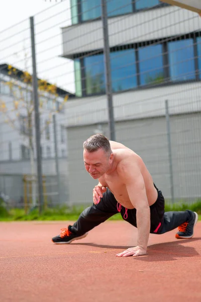 Shirtless middle age man doing one arm push ups on a sports field