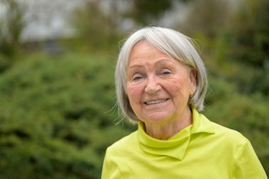 80 'li yaşlarda, beyaz gri saçlı ve yeşil gömlekli yaşlı bir kadın kameraya gülümsüyor.