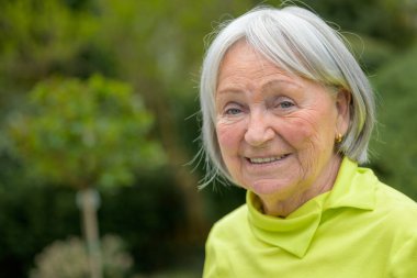 80 'li yaşlarda, beyaz gri saçlı ve yeşil gömlekli yaşlı bir kadın kameraya fotokopi aletiyle gülümsüyor.