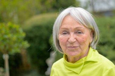 80 'li yaşların başında, beyaz-gri saçlı ve yeşil gömlekli yaşlı bir kadın kameraya sıkılmış gibi görünüyor.