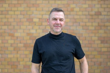 Orta yaşlı, gri saçlı bir adam, tuğla bir duvarın önündeki kameraya arkadaş gibi bakıyor.