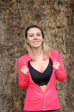 Sarışın kadın parkta bir ağacın önünde duruyor ve kamerayla eğleniyor.