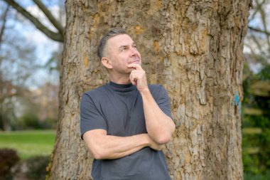 Düşünceli, kır saçlı bir adam parktaki bir ağacın önünde dikiliyor ve yukarı bakıyor.