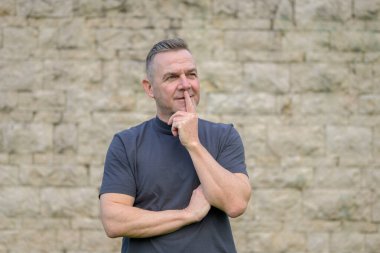 Gri saçlı bir adam duvarın önünde dikiliyor ve düşünceli bir şekilde yan bakıyor.