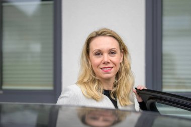 Orta yaşlı bir kadın arabaya binerken kameraya bakar.