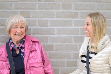 Anne ve kızı yürekten gülerken beyaz bir duvarın önünde duruyorlar.