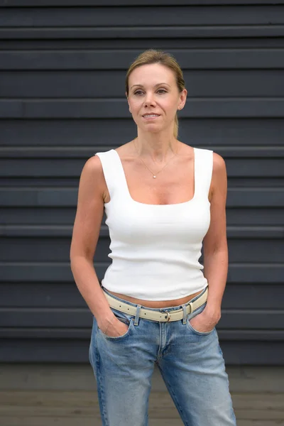stock image Three quarter length portrait of an attractive forty year old woman wearing a white top and jeans with her hands in her pockets smiling friendly looking to the side