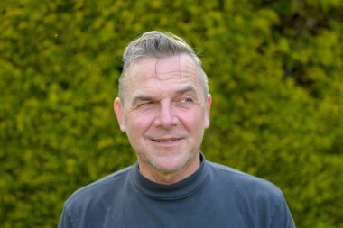 Bahçede duran, beyaz saçlı, orta yaşlı bir adamın portresi.