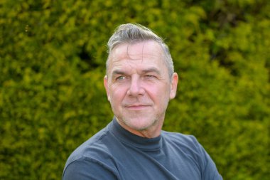 Orta yaşlı bir adamın portresi. Gri saçlı. Bahçede duruyor ve tarafsız görünüyor.