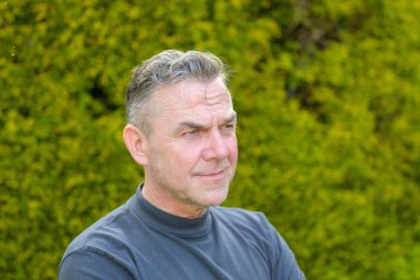 Bahçede duran, beyaz saçlı, orta yaşlı bir adamın portresi.