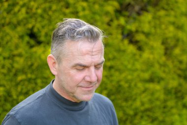 Bahçede duran, beyaz saçlı, orta yaşlı bir adamın portresi.