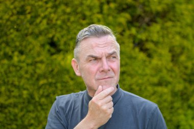 Bahçede duran gri saçlı, orta yaşlı bir adamın portresi. Parmağını çenesine dayamış şekilde aşağıya bakıyor.