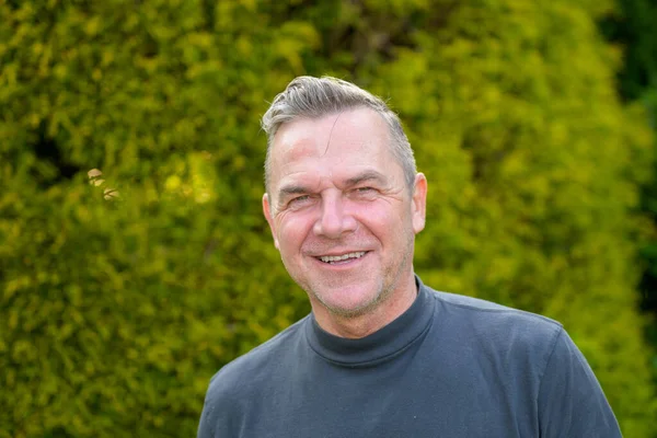 Bahçede duran ve kameraya dost canlısı bakan orta yaşlı bir adamın portresi.