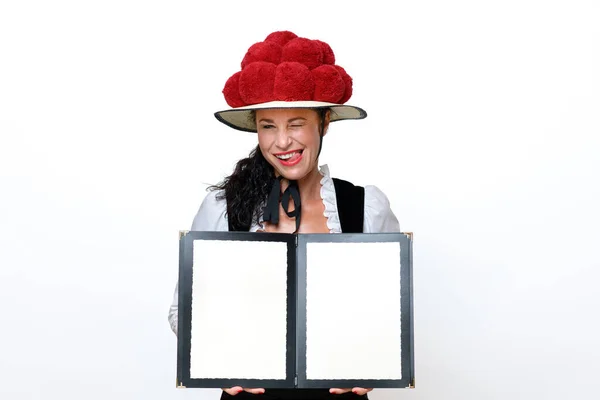 在一个有着红色华丽花朵的传统的宝莲小屋里当黑森林女服务员 她调皮地眨着眼睛 伸出舌头 带着友好的微笑 开着一个空白的菜单 — 图库照片