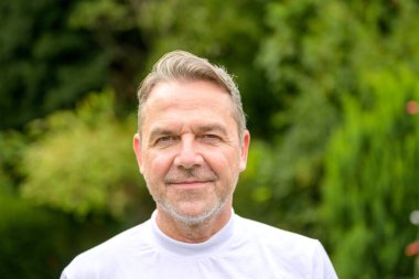 50 'li yaşlarda çekici, orta yaşlı bir adamın portresi. Bahçede dikilirken yüzünde hafif bir gülümsemeyle.