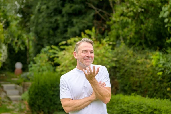 stock image Attractive middle aged man in his 50s smiling and making a gesture of doubt and questionability while standing in the garden