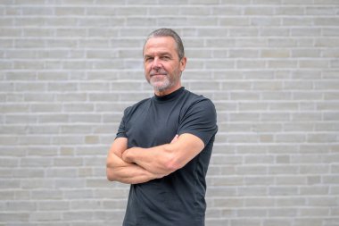 Side view of an attractive man with crossed arms looking to the camera in a friendly and determined manner in front of a white brick wall clipart