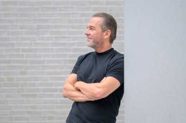 Gri saçlı çekici bir adam, kolları çapraz bir şekilde duvara dayanıyor ve beyaz tuğla bir duvarın önündeki mesafeye umutla bakıyor.