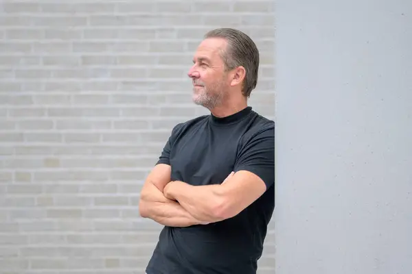 Gray haired attractive man leans against a wall with his arms crossed and looks expectantly into the distance in front of a white brick wall