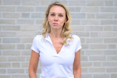 Beyaz tuğla bir duvarın önünde kameraya bakan çekici sarışın bir kadının üst vücut portresi.