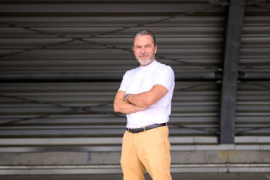 Beyaz tişörtlü, çekici, gri saçlı bir adamın dörtte üçlük vücut portresi kollarını kavuşturmuş, şık bir saat takıyor ve kıvrımlı demir bir duvarın önünde kameraya samimi bir şekilde bakıyor.