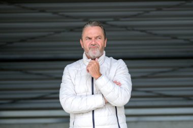 Çekici, gri saçlı, ceketinde beyaz bir kışlık ceket olan, elleri ceketinin üzerinde, bükülmüş demir bir duvarın önünde huysuz görünen bir adam.