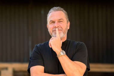 Düşünceli, beyaz saçlı, siyah t-shirt giyen bir adamın parmakları dudaklarında, tahta bir duvarın önünde.