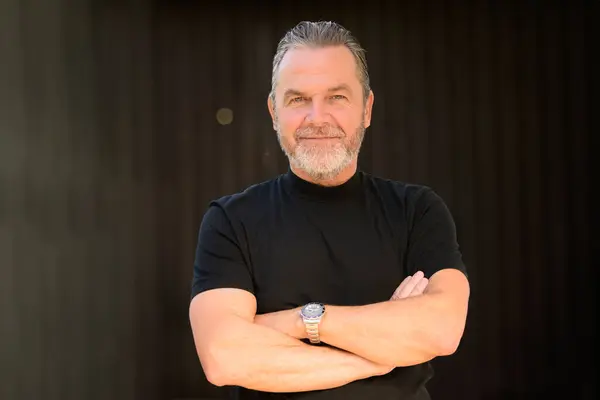 stock image Attractive gray-haired man wearing a black T-shirt and wearing a stylish watch and looks friendly to the camera, in front of a wooden wall