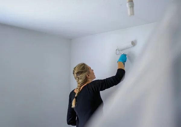 Back view of a woman painting the wall of her apartment with a large paint roller, in her new apartment