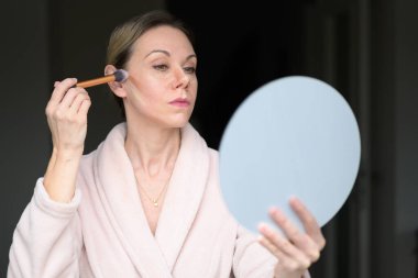 Sarışın kadın fırçayla makyaj yapıyor ve evde aynaya bakıyor.