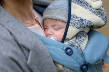 Annesinin göğsünde ya da göğüs dekoltesinde uyuyan bir bebeğin, mavi ve beyaz çizgili bir şapka takarak son derece samimi bir yakınlığı.